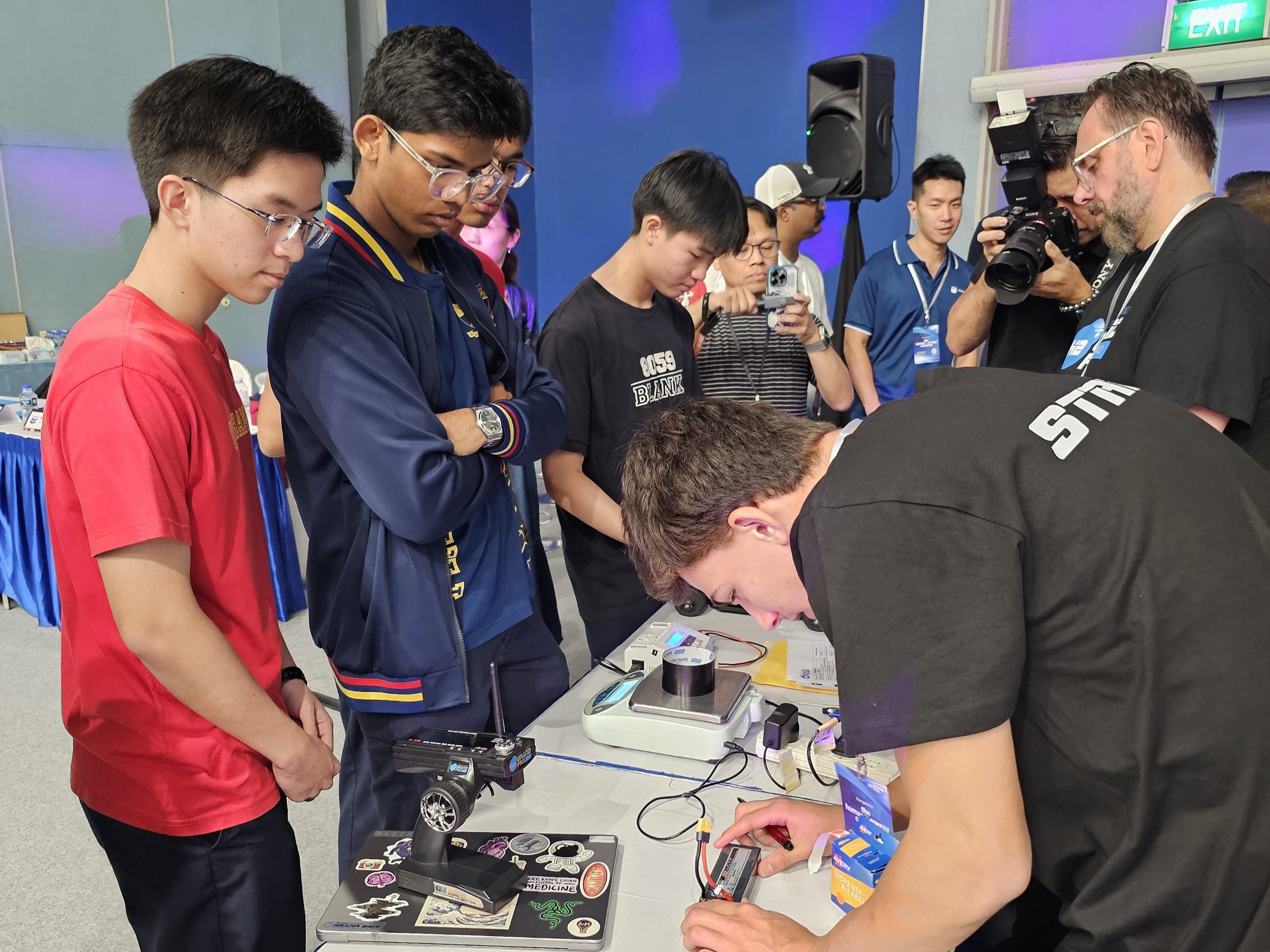 A group of young people examines an interactive tech demonstration. One person is adjusting equipment on a table filled with gadgets, creating a focused learning atmosphere.